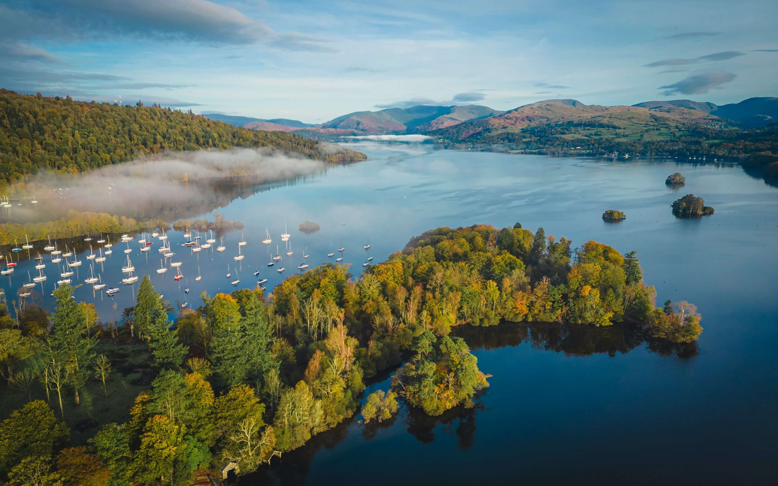 A lake with wooded islands, moored boats and mist beneath surrounding hills